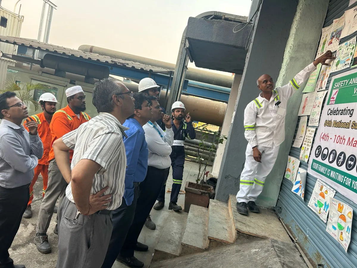 Personnel in hard hats and work clothes listen to a speaker beside pipework and a safety display at a terminal.