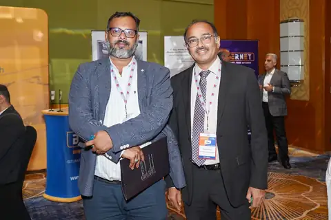 Two suited delegates wearing event lanyards pose together beside a conference exhibition area.