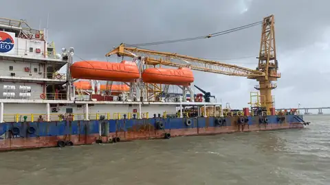 The side of a crane vessel shows orange enclosed lifeboats, white accommodation decks and a yellow crane above a blue hull.
