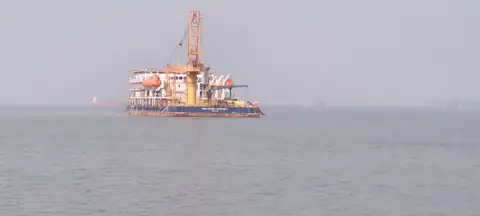 A distant crane vessel with a white accommodation block is seen through haze across open water.