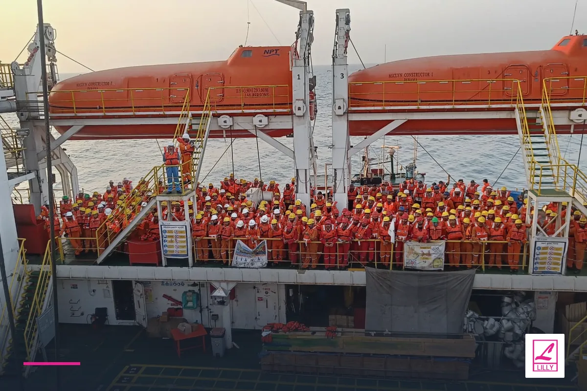 Crew in orange lifejackets assemble beneath orange enclosed lifeboats, with a navy and magenta Lilly Maritime treatment.